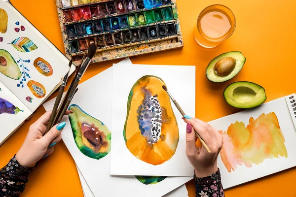 Вид сверху женских рук, рисующих авокадо и папайю акварельными красками, кисть на желтом столе — стоковое фото