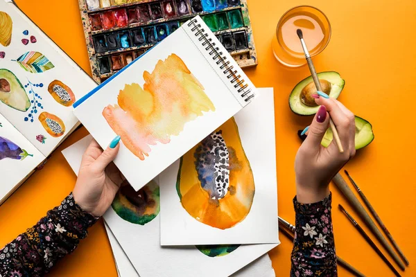 Vista superior de las manos femeninas dibujando aguacate y papaya con pinturas de acuarela, pincel sobre mesa amarilla - foto de stock