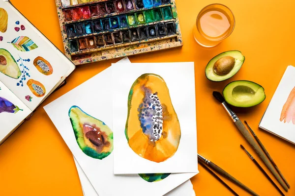 Vista superior de los dibujos de aguacate y papaya junto a pinturas de colores, pinceles sobre fondo amarillo - foto de stock