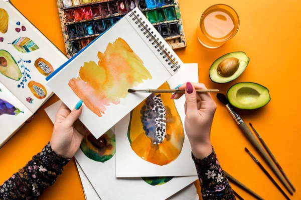 Top view of female hands drawing papaya with watercolor paints and paintbrush on yellow table — Stock Photo