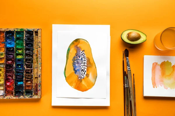 Top view of album with papaya drawing next to colored paints and paintbrushes   on yellow background — Stock Photo