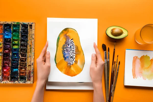 Vista superior de las manos femeninas sosteniendo la imagen de papaya sobre la mesa amarilla con utensilios de dibujo - foto de stock