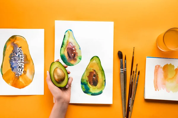 Вид сверху на женскую руку, держащую авокадо над желтым столом с рисунками фруктов и кистей — стоковое фото