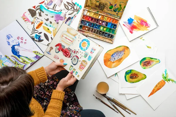 Vista superior de la mujer sosteniendo álbum rodeado de dibujos a color y utensilios de dibujo — Stock Photo