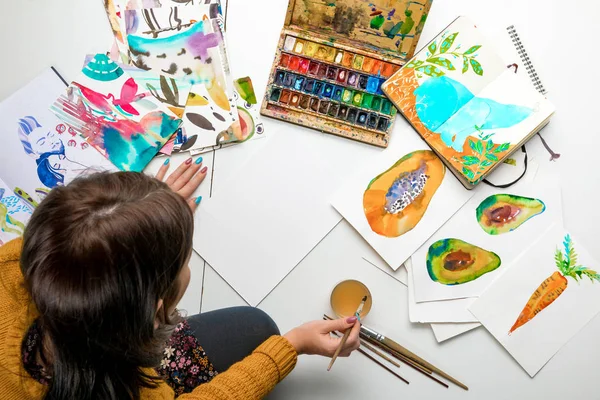 Vista superior de la mujer que se prepara para pintar con acuarelas pinturas mientras está rodeado de dibujos a color y utensilios de dibujo - foto de stock