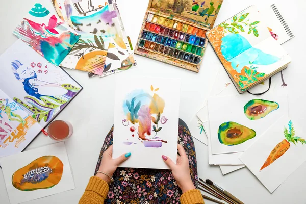 Top view of woman holding watercolor drawing while surrounded by color pictures — Stock Photo
