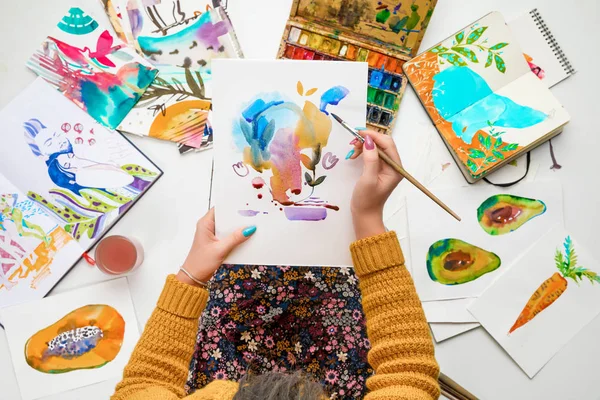 Draufsicht einer Frau, die eine Zeichnung auf Knien hält und sie mit Aquarellfarben bemalt, während sie von farbigen Bildern umgeben ist — Stockfoto