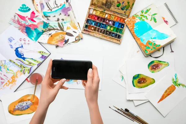 Enfoque selectivo de manos femeninas haciendo foto de pinturas de acuarela y utensilios de dibujo usando teléfono inteligente - foto de stock