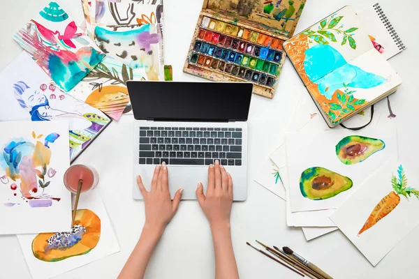 Vista superior de las manos femeninas en el ordenador portátil mientras está rodeado de dibujos de acuarela - foto de stock