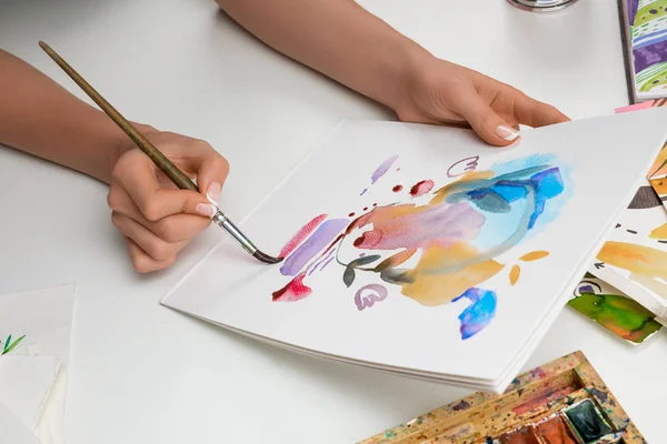 Enfoque selectivo de las manos femeninas pintura con acuarelas pinturas y pincel sobre papel blanco - foto de stock