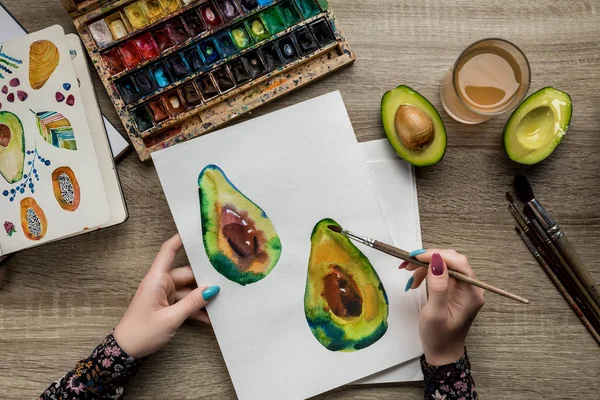 Vista superior de mãos femininas desenho de abacates com tintas aquarela e pincel em mesa de madeira — Fotografia de Stock