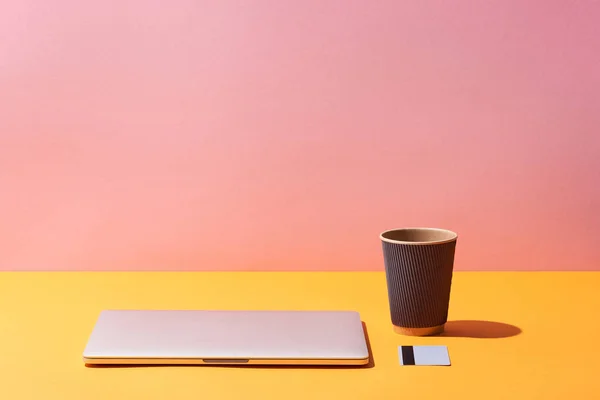 Kaffeetasse aus Papier in der Nähe von Laptop und Kreditkarte auf gelber Oberfläche und rosa Hintergrund — Stockfoto