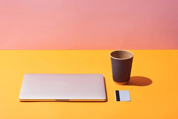 Paper coffee cup near laptop and credit card on yellow surface and pink background — Stock Photo