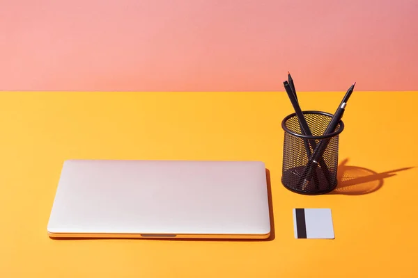 Porta-lápis perto de laptop e cartão de crédito na superfície amarela e fundo rosa — Fotografia de Stock