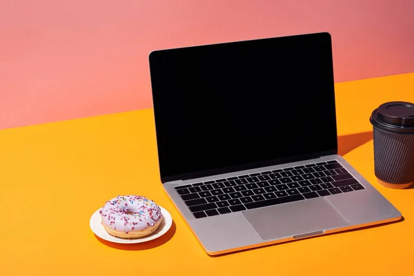 Portátil con pantalla en blanco cerca de rosquilla y taza de café de papel en la superficie amarilla y fondo rosa - foto de stock