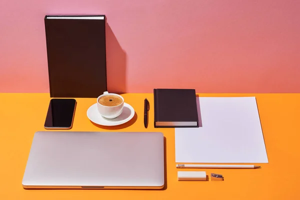Notebooks, laptop, smartphone, taza de café, platillo, bolígrafo, basura, borrador y hoja de papel sobre escritorio amarillo y fondo rosa - foto de stock