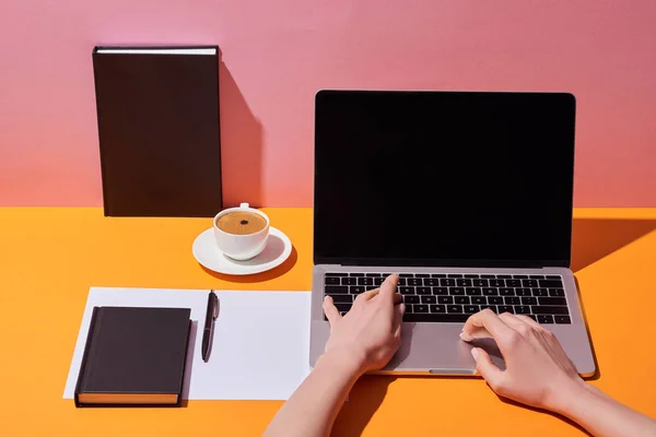 Обрізаний вид жінки з ноутбуком з порожнім екраном біля чашки кави, блюдця, ручки, паперового листа та блокнотів на жовтому столі та рожевому фоні — стокове фото