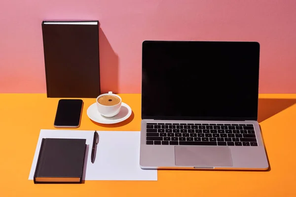 Laptop with blank screen near coffee cup, saucer, pen, paper sheet and notebooks on yellow desk and pink background — Stock Photo