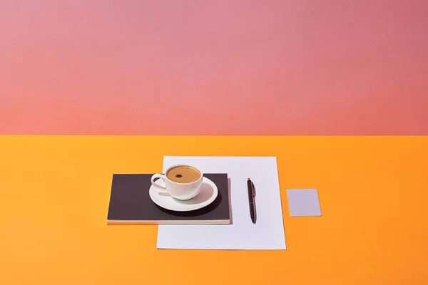 Xícara de café, pires, caneta, folha de papel e caderno na mesa amarela e fundo rosa — Fotografia de Stock
