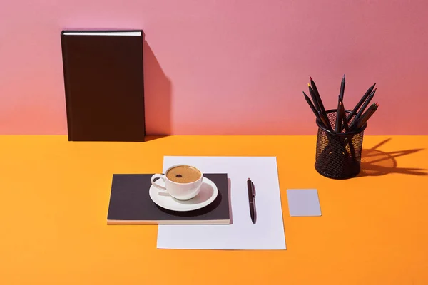 Xícara de café, pires, caneta, folha de papel, suporte de lápis e notebook na mesa amarela e fundo rosa — Fotografia de Stock
