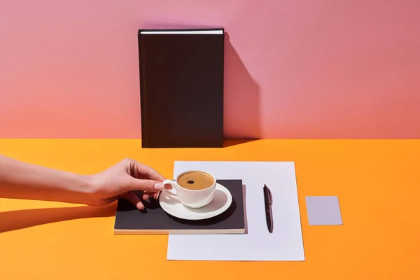 Vista recortada de la mujer sosteniendo taza de café cerca de platillo, pluma, hoja de papel y cuadernos sobre escritorio amarillo y fondo rosa - foto de stock