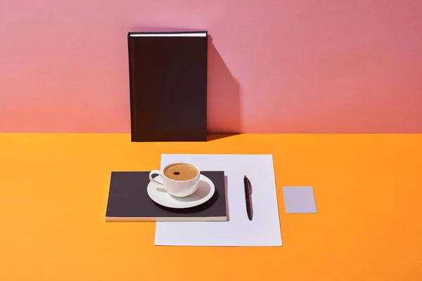 Tazza di caffè vicino piattino, penna, foglio di carta e quaderni su scrivania gialla e sfondo rosa — Foto stock