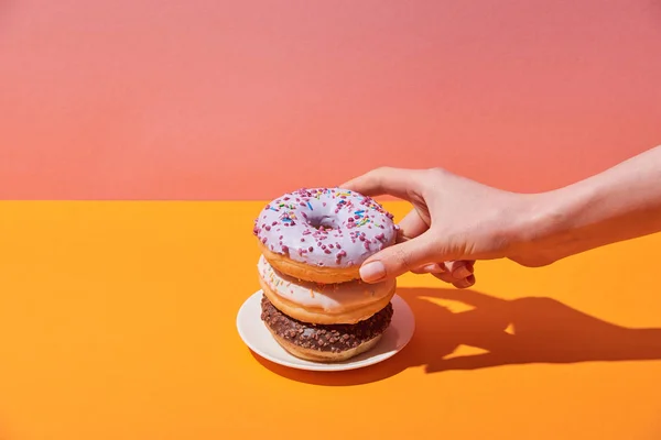 Vista cortada de mulher tomando saborosos donuts no pires na mesa amarela e fundo rosa — Fotografia de Stock