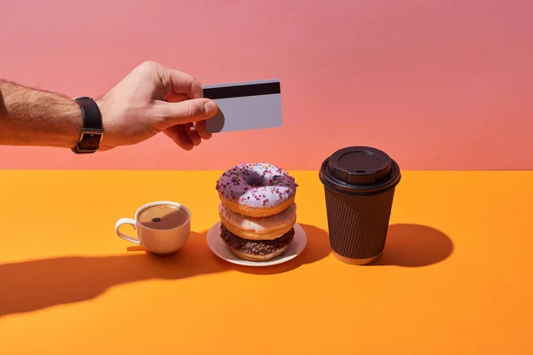 Vista cortada do homem peying para saborosos donuts no pires e xícaras de café na mesa amarela e fundo rosa — Fotografia de Stock