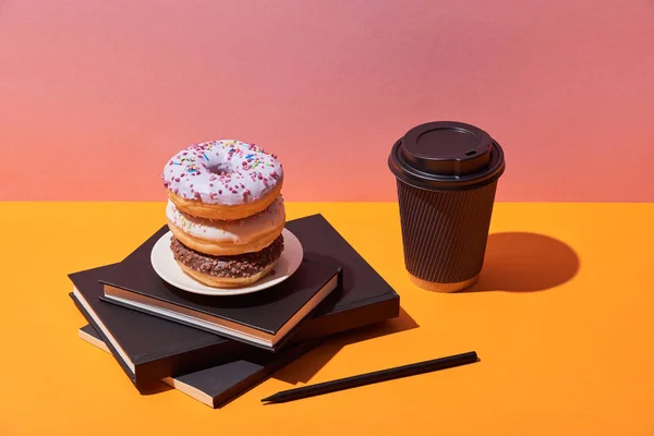 Saborosos donuts no pires e xícara de café de papel perto de cadernos e lápis na mesa amarela e fundo rosa — Fotografia de Stock