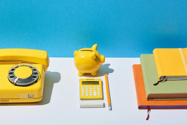 Multicolored notebooks, calculator, telephone and piggy bank on white desk and blue background — Stock Photo