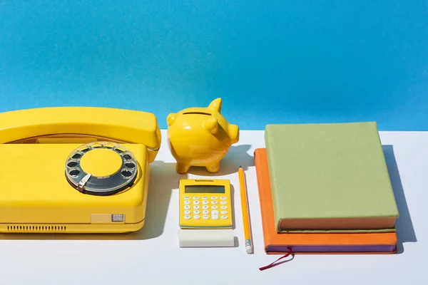 Quaderni multicolore, calcolatrice, telefono e salvadanaio su scrivania bianca e sfondo blu — Foto stock