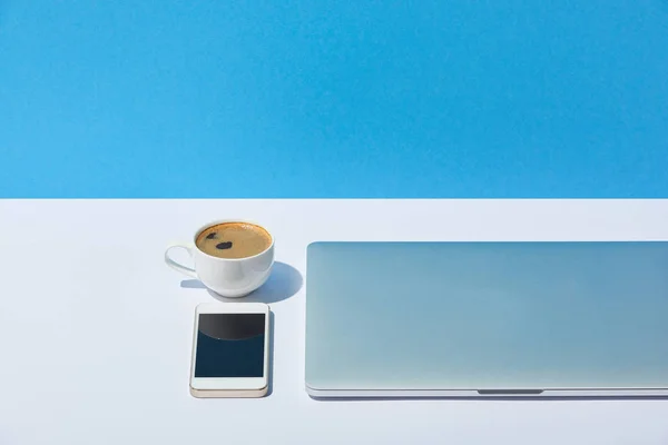 Laptop, coffee cup and smartphone on white desk and blue background — Stock Photo