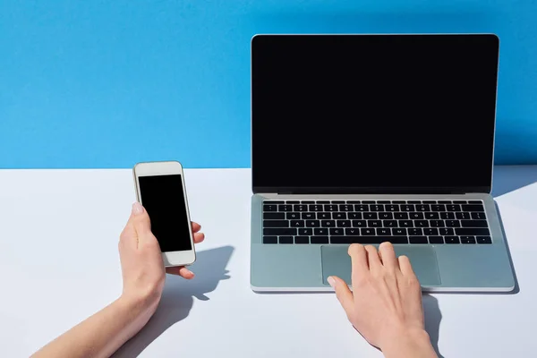 Vue recadrée des femmes utilisant un ordinateur portable avec écran vierge et tenant smartphone sur fond blanc et bleu — Photo de stock
