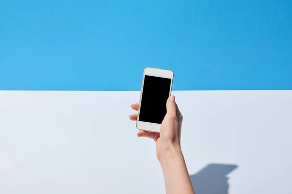 Vista recortada de la mujer usando teléfono inteligente con pantalla en blanco sobre escritorio blanco y fondo azul - foto de stock