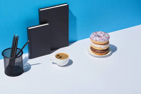 Tasty donuts, coffee cup, pencil holder and notebooks on white desk and blue background — Stock Photo