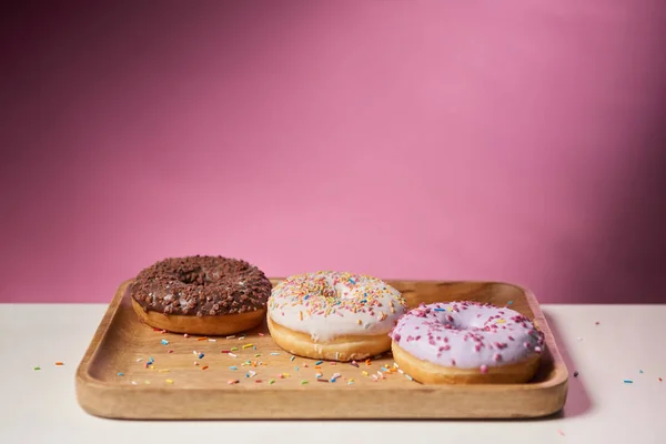 Вкусные глазурованные пончики посыпаются на деревянную доску для резки на розовом фоне — стоковое фото