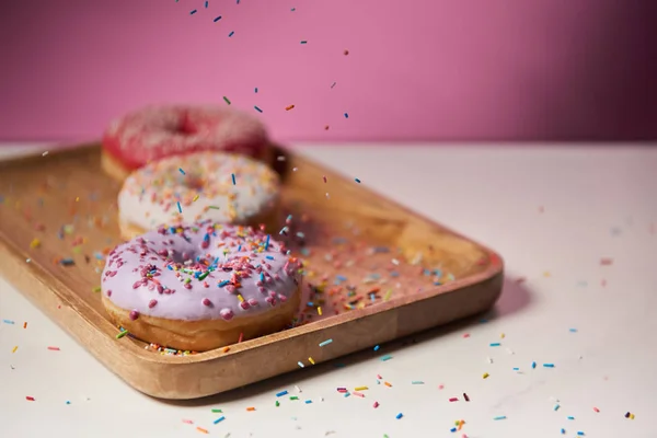 Selektiver Fokus von leckeren Donuts mit Zuckerguss und fallenden Streusel auf dem Holzschneidebrett — Stockfoto
