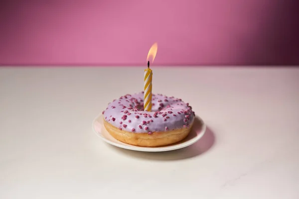 Burning candle in middle of doughnut with sprinkles on white table on pink background — Stock Photo