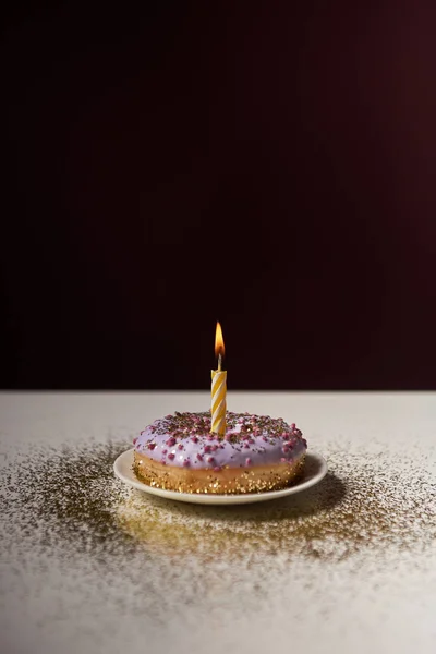 Burning candle in middle of glazed doughnut with golden sparkles on white table isolated on black — Stock Photo