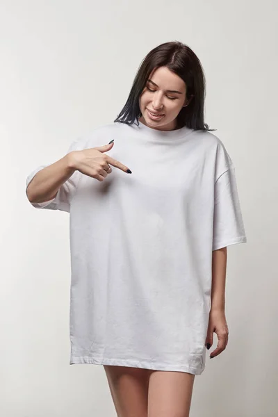 Beautiful young woman pointing with finger at white oversize t-shirt with copy space isolated on grey — Stock Photo