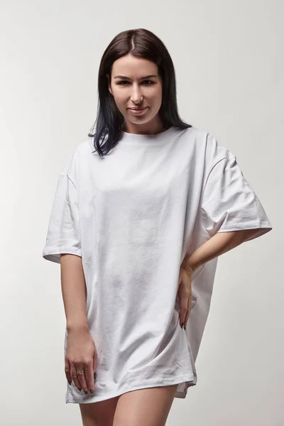 Belle jeune femme en t-shirt oversize blanc avec espace de copie regardant la caméra isolée sur gris — Photo de stock