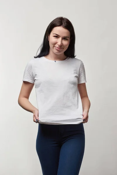 Hermosa mujer joven sonriente en camiseta blanca con espacio de copia mirando a la cámara aislada en gris - foto de stock