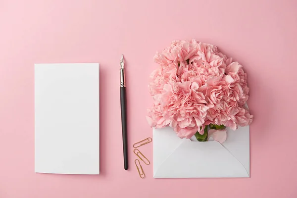 Draufsicht auf Blankokarte, Federhalter und rosa Blumen in weißem Umschlag isoliert auf rosa — Stockfoto