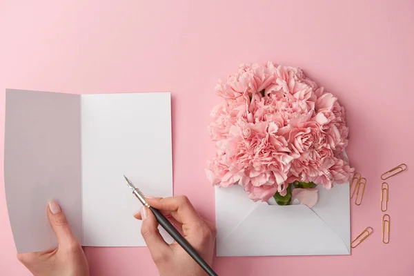 Recortado tiro de mujer escribiendo en tarjeta de felicitación y flores de color rosa en sobre aislado en rosa - foto de stock