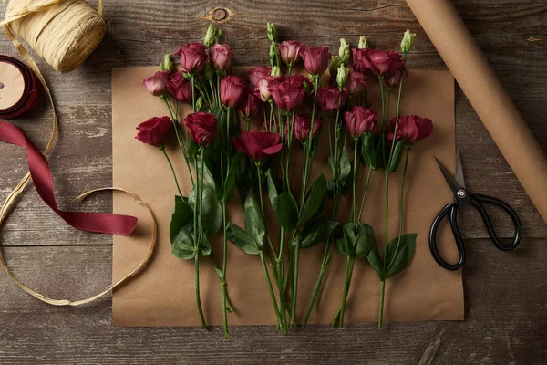 Vista superior de belas flores de eustoma vermelho em papel artesanal, tesouras e fitas na superfície de madeira — Fotografia de Stock