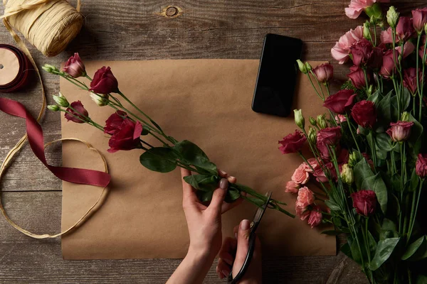 Cropped shot of woman cutting flowers above wooden table with craft paper and smartphone — Stock Photo