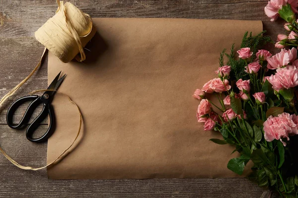 Vista superior de hermosas flores rosadas, tijeras, cinta y papel artesanal en la superficie de madera - foto de stock