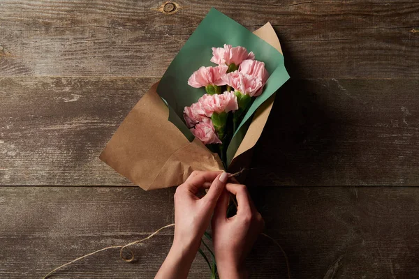 Cropped shot of woman wrapping pink carnation flowers in craft paper on wooden surface — Stock Photo