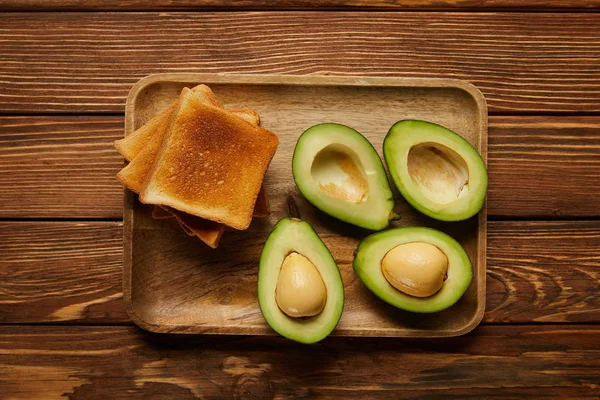 Draufsicht auf geschnittene Avocados und gegrillte knusprige Toasts auf Holzgrund — Stockfoto