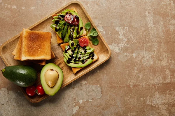 Top view of toasts with avocados, cherry tomatoes on wooden chopping board,  brown background — Stock Photo